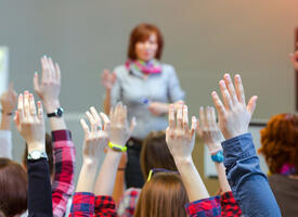 A teacher stands in front of a class with many raised hands