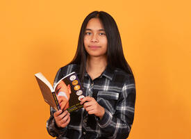 Boy with long dark hair wearing a black plaid shirt reading a book infront of an orange wall