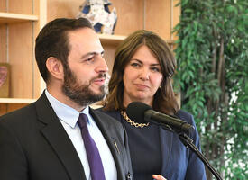 Man with a beard speaks at a microphone with a women standing behind him in a navy suit