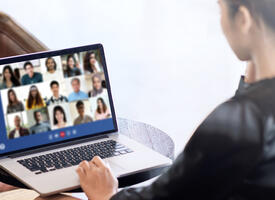 A person wearing a black blouse sitting in front of a laptop screen that has 15 faces on it, all people part of an online meeting