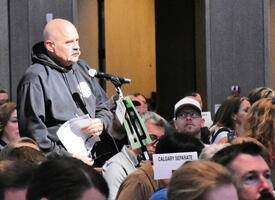 Bald man in a grey hoodie standing in a crowd at a microphone