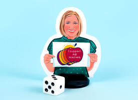 An illustrated boardgame player piece sits on a blue background in front of a die. The illustration of Melanie shows her with a blonde bob hairstyle, wearing a green short-sleeved shirt, holding a "I support AB teachers" graphic.