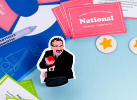 A player piece of Mark Ramsankar sits amongst other boardgame materials. Mark Ramsankar is illustrated holding a red apple and wearing a maple leaf tie. There are tokens with gold stars near him, and the board game has an illustration of Canada's parliament building.