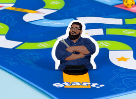 A photograph of a bright blue boardgame with a focus on a player token at the start of the game. It is an illustration of D'Souza in his navy blue mechanic suit, holding a wrench and smiling.