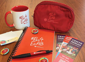 A Red4EdAB branded notebook, coiled pencil case, and mug sit on a wooden table with three bookmarks, three "I support AB teachers" button pins, red ATA branded pen, and two small notepads with "I support AB teachers and ATA branding.