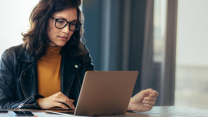 Woman in leather jacket and yellow shirt looking at laptop next to a window
