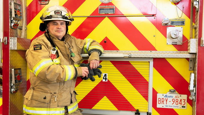 Firefighter standing in full uniform leaning on the back of a fire truck with a high contrast yellow and red chevron pattern.