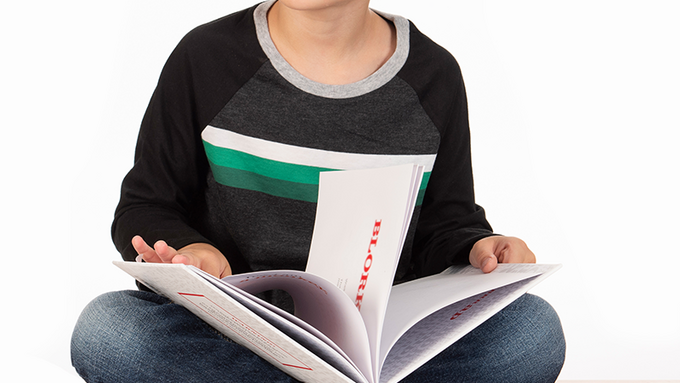 White boy with baseball shirt sits cross-legged on a table with a book in his lap.