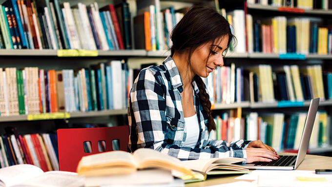 Young women of colour sitting at a table in a library writing with a pencil in front of an open laptop. 