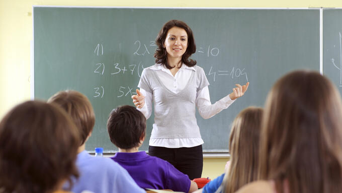 White female with brown hair standing in front of a chalkboard teaching students in the foreground 