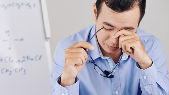 Middle age man rubbing holding his glasses and rubbing his eyes infront of a white board with equations