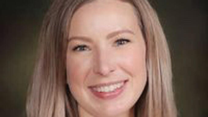 Portrait of a smiling white women with blonde hair in a black blazer and white blouse in front of a dark background. 
