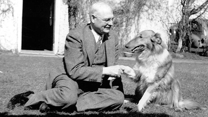 black and white photo of a caucasian man seated on the ground in a suite, shaking the paw of a rough collie dog.