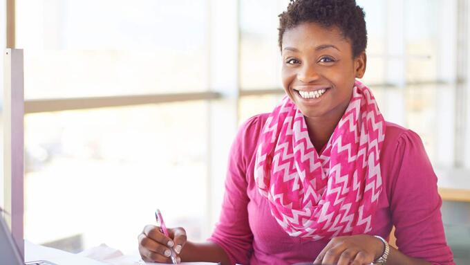 Women with short black hair wearing a fuscia scarf and sweater smiling while working at a desk.