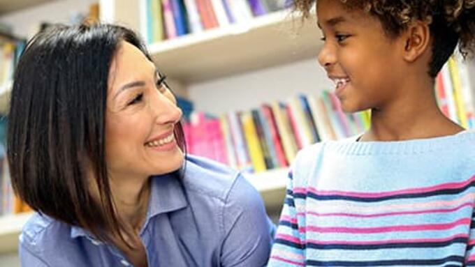 Teacher in blue shirt seated next to a young male student in stiped purple shirt smiling at each other