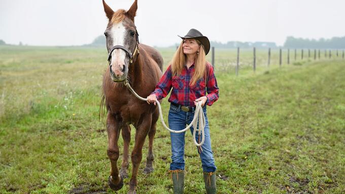 Women in a cowboy had standing in a field leading a chestnut coloured horse