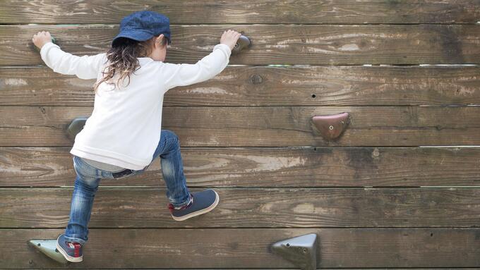 Young child climbing a wall with grips wearing blue jeans, a hat and white shirt