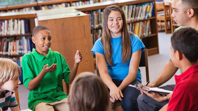 Students and their teacher seated in chairs listening to a young boy speak