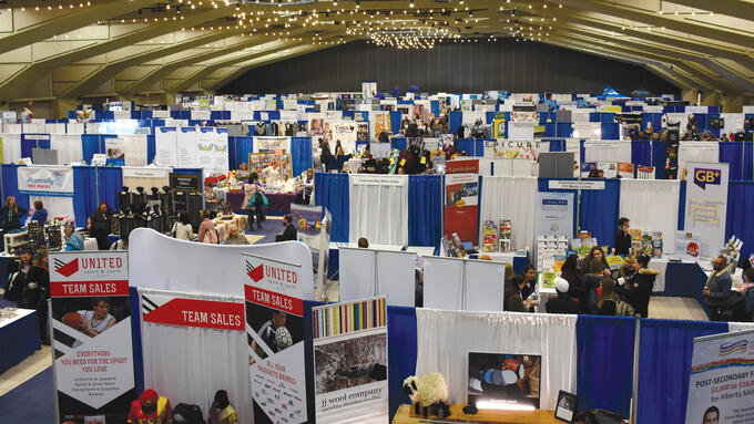 A photo of the inside of a large convention centre. The top of the photo is yellow with beams of the roof and lights. On the floor are people, many displays and blue dividers for the booths.