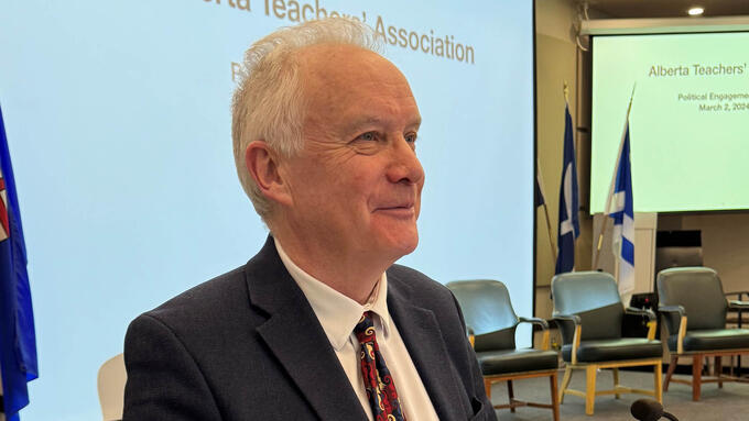 Grey haired man stands at a podium in front of an audience.