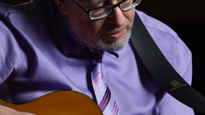 A person in glasses and a purple shirt and tie playing a guitar