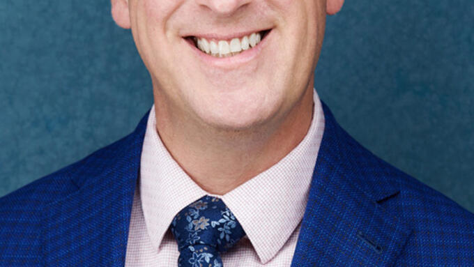 A white man in a blue suit. The man is wearing glasses and is standing in front of a lighter blue background. 