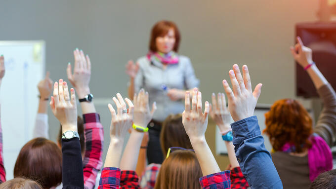 A teacher stands in front of a class with many raised hands