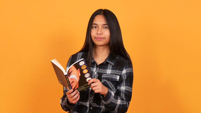 Boy with long dark hair wearing a black plaid shirt reading a book infront of an orange wall
