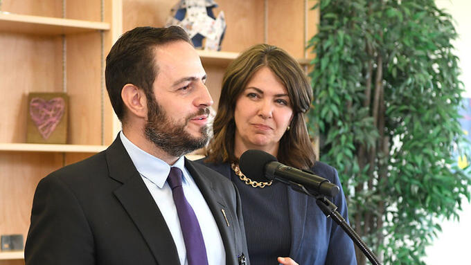 Man with a beard speaks at a microphone with a women standing behind him in a navy suit