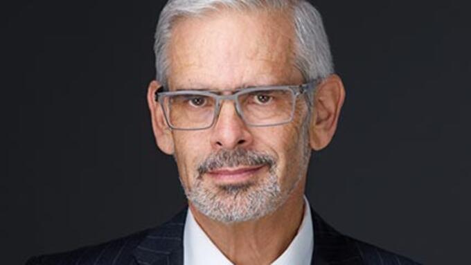 Grey haired man in silver glasses wears a dark suit infront of black background 