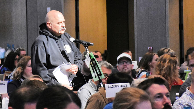 Bald man in a grey hoodie standing in a crowd at a microphone