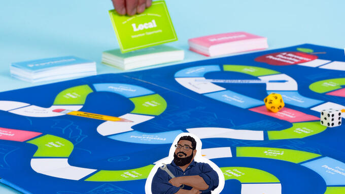 A photograph of a bright blue boardgame on a light blue background. In the background, a man's hand pick up playing cards that say "Local volunteer opportunity". In the foreground, a player token is at the start of the boardgame. The player is an illustration of D'Souza in his mechanic outfit, crossing his arms confidently and holding a wrench.