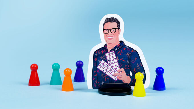 An illustrated boardgame player piece of Sandy Johnson stands among a colourful collection of more generic conical player pieces. Sandy is wearing glasses, smiling, and holding up a math game with numbers in a chart..
