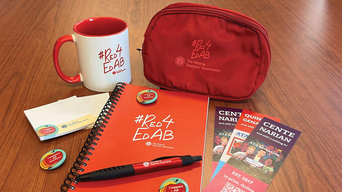 A Red4EdAB branded notebook, coiled pencil case, and mug sit on a wooden table with three bookmarks, three "I support AB teachers" button pins, red ATA branded pen, and two small notepads with "I support AB teachers and ATA branding.