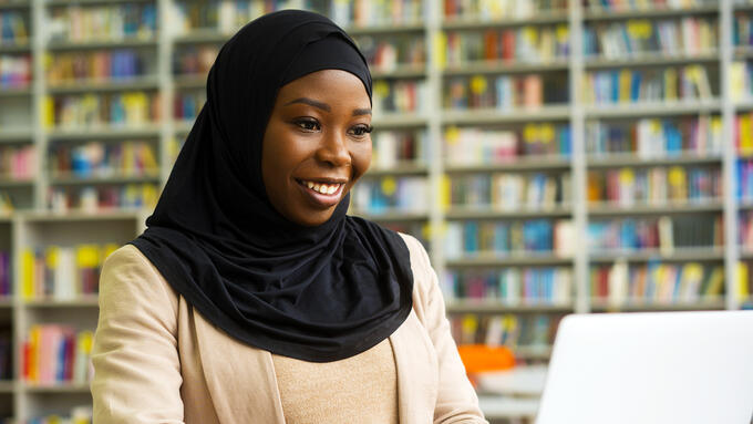 A woman in a hijab using a laptop in a library. 