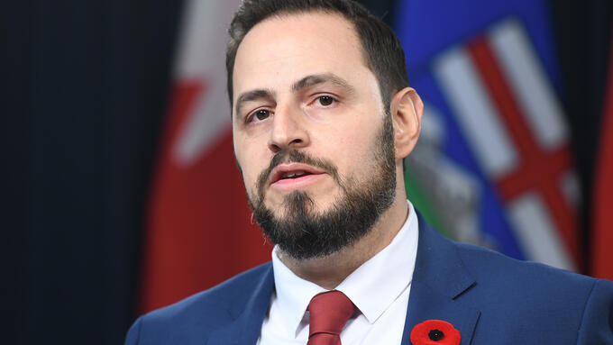 Education Minister infront of flags wearing a poppy in a blue suit