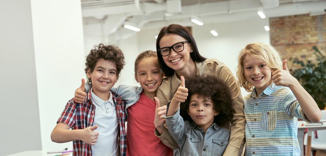 Teacher wearing glasses standing with 4 children all giving a thumbs up