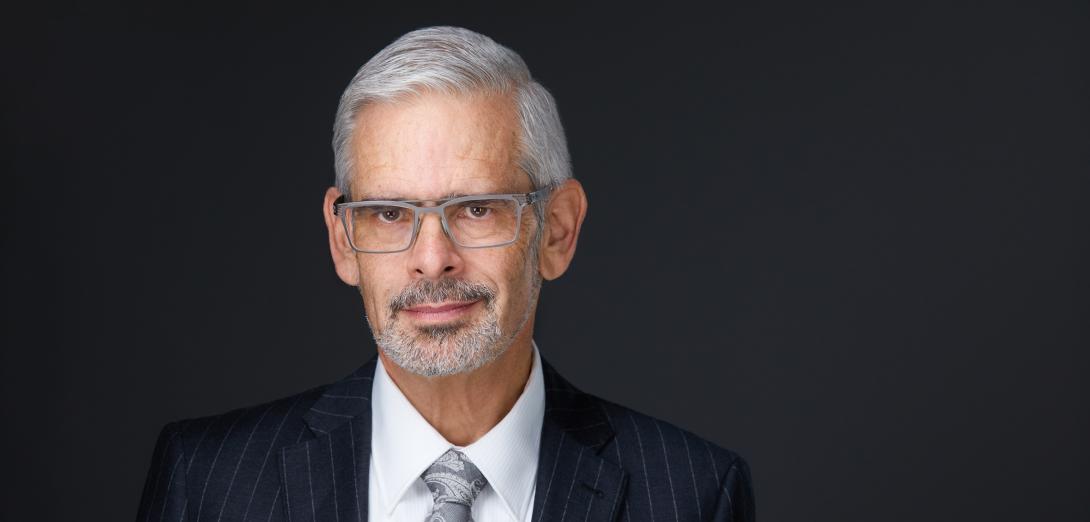 Dennis Theobald with silver white goatee and hair wearing a dark suit and tie in front of a black background.