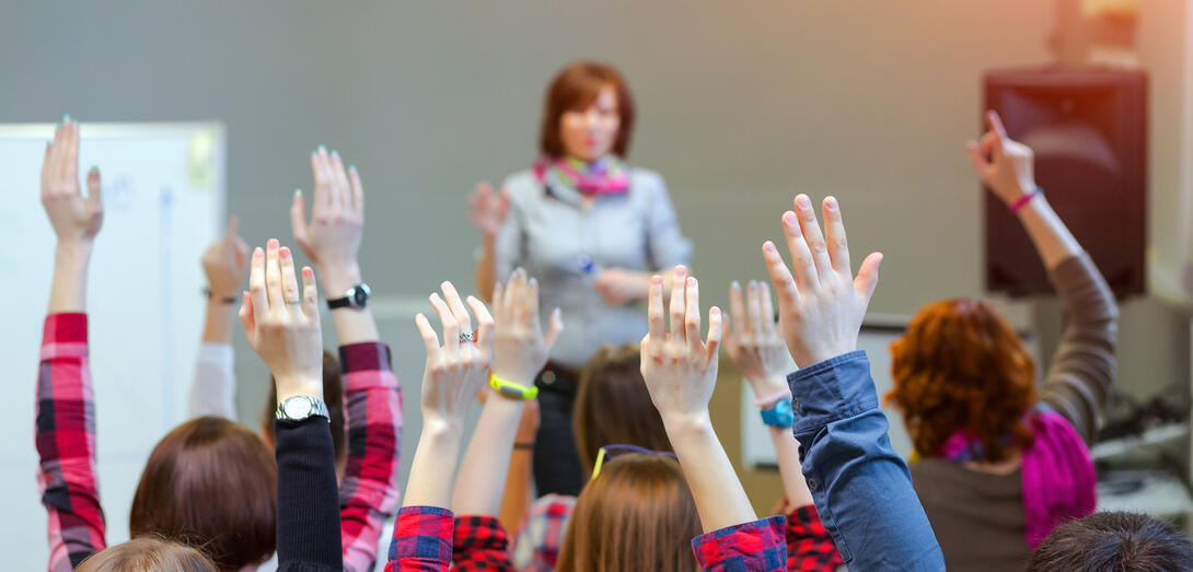 A teacher stands in front of a class with many raised hands