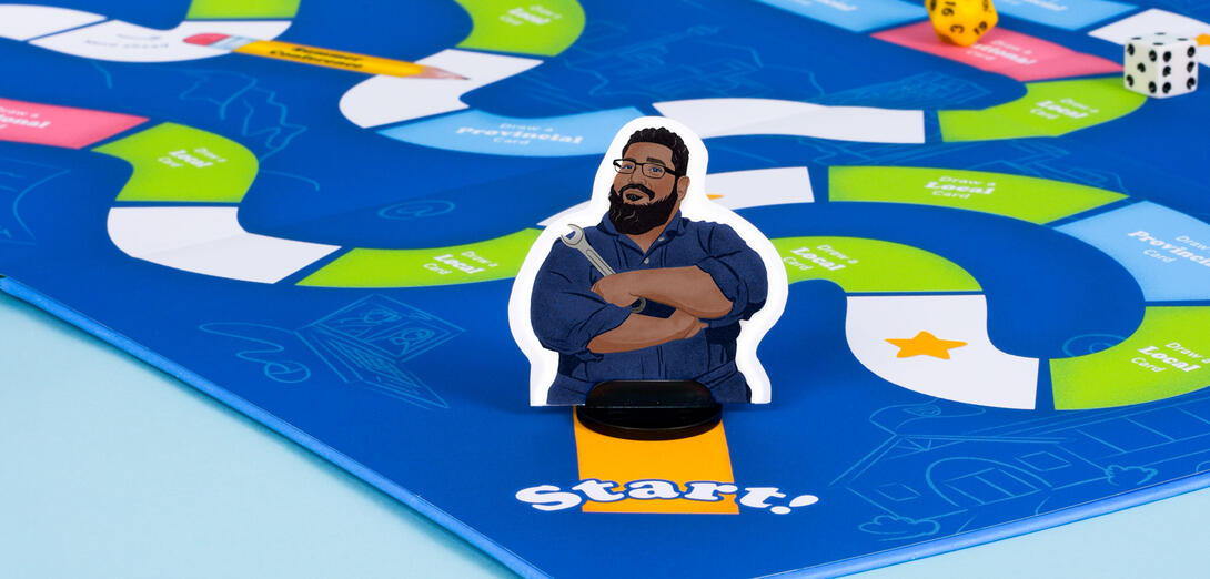 A photograph of a bright blue boardgame with a focus on a player token at the start of the game. It is an illustration of D'Souza in his navy blue mechanic suit, holding a wrench and smiling.