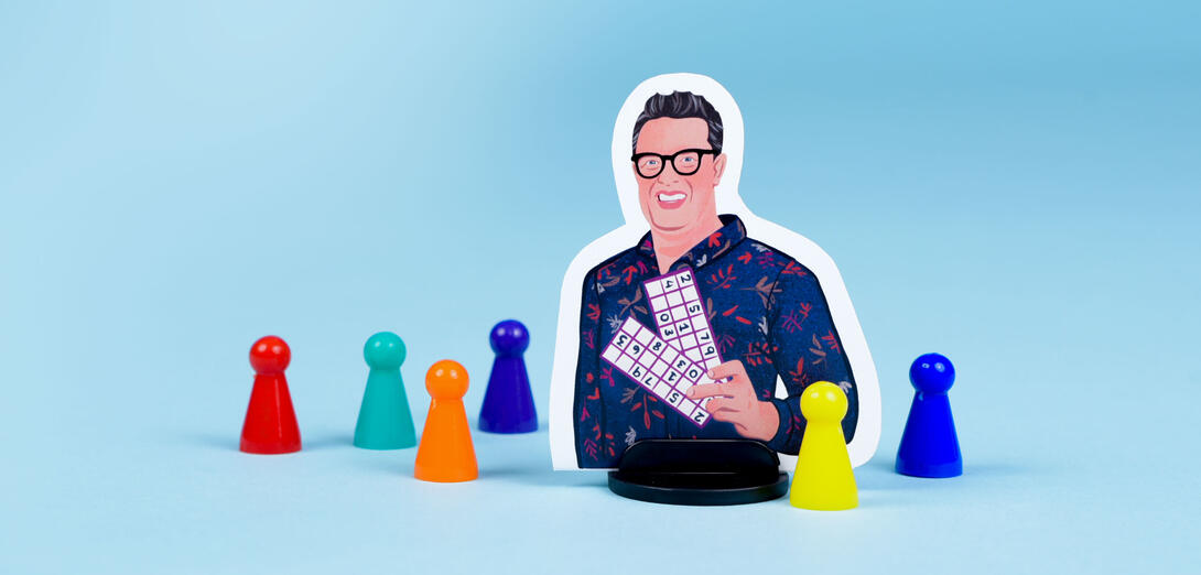 An illustrated boardgame player piece of Sandy Johnson stands among a colourful collection of more generic conical player pieces. Sandy is wearing glasses, smiling, and holding up a math game with numbers in a chart..