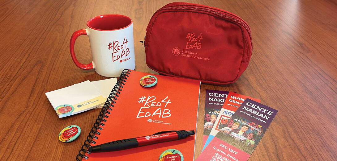 A Red4EdAB branded notebook, coiled pencil case, and mug sit on a wooden table with three bookmarks, three "I support AB teachers" button pins, red ATA branded pen, and two small notepads with "I support AB teachers and ATA branding.